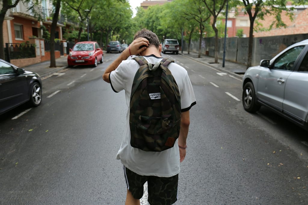 Protecting Your Child's Inheritance from Addiction and Debt A young person wearing a camouflage backpack and shorts stands in the middle of a street, looking down with their hand on their head in a gesture of stress or confusion.