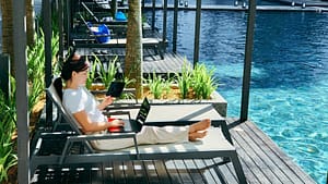 The Digital Nomad Lifestyle: Why Your Legal Residency Matters for Estate Planning A woman working on a laptop and holding a tablet while relaxing on a lounge chair by a swimming pool, illustrating the digital nomad lifestyle.