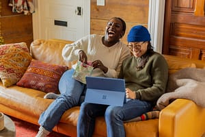 Planning for Your Family's Future: Estate Planning and Living Trusts Two women laughing while sitting on a leather couch, one opening a gift bag and the other using a Microsoft Surface tablet.