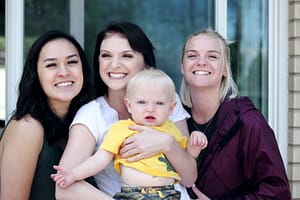 Protecting Your Family: Estate Planning for Same-Sex Couples Three smiling women posing together outdoors, with one woman holding a young blonde toddler in a yellow shirt and camouflage shorts.